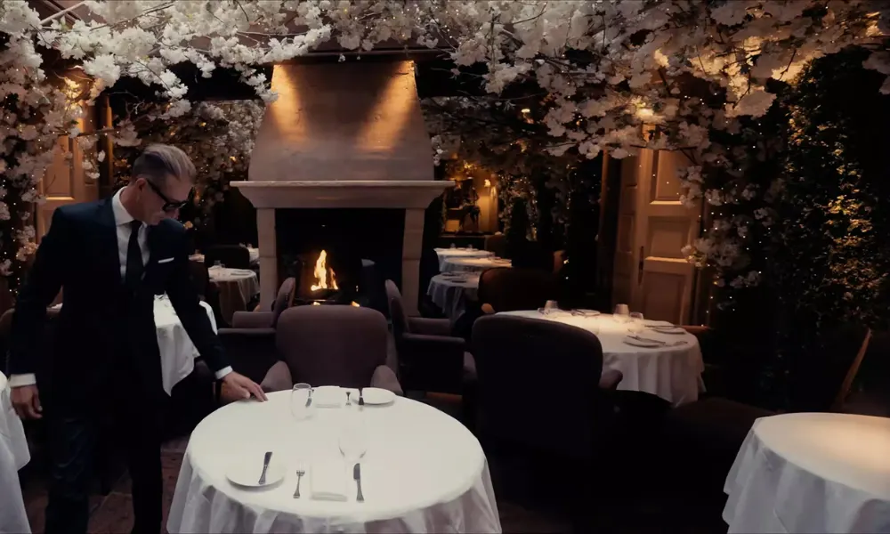 A waiter sets tables with floral displays and fire behind
