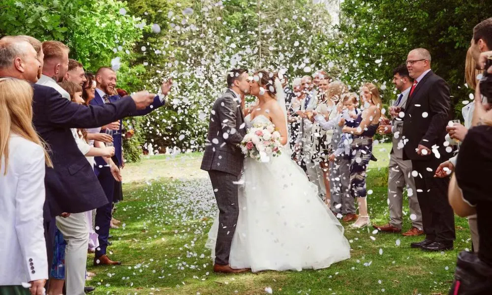 a bride and groom kiss as crowds throw confetti in a woodland setting