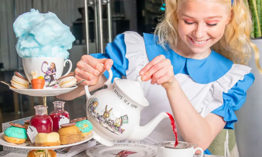 Girl in Alice costume pours pink tea behind tiers of cake