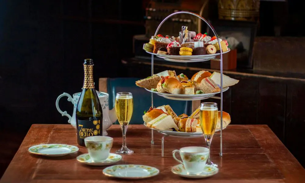 A tea plate full of sandwiches and cakes with cups, saucers, a bottle of Prosecco and two glasses