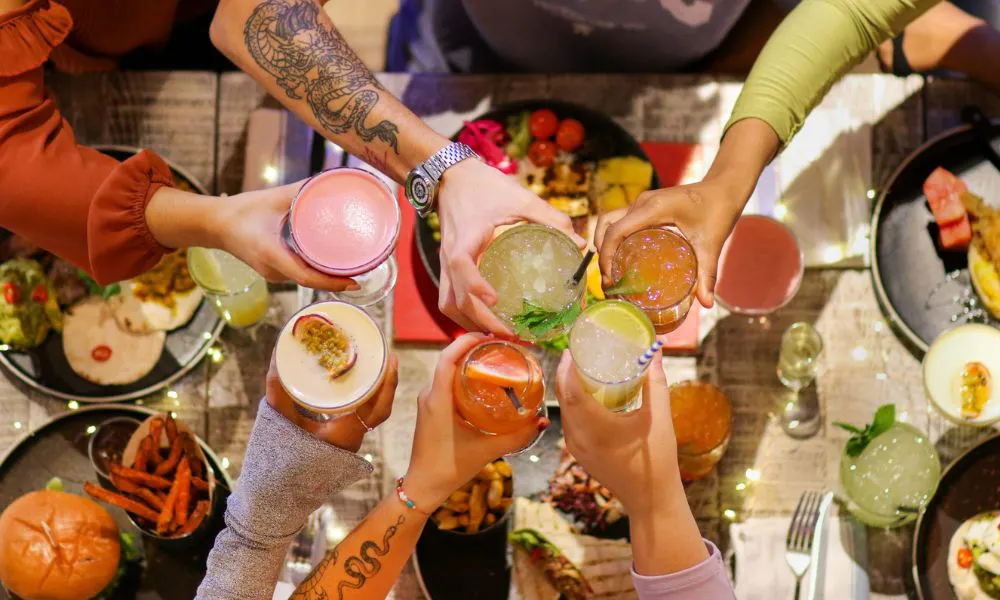 A group of people cheersing with cocktails over a table which is full of different dishes and sides including sweet potato fries 